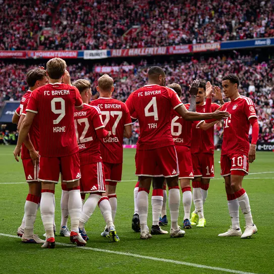 A group of FC Bayern Munich players in red jerseys huddle together on the field, celebrating and discussing strategies. The crowd is visible in the background, creating an energetic and vibrant atmosphere.