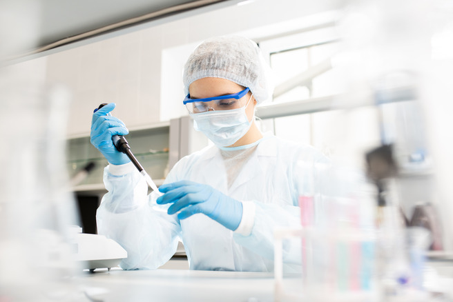 A laboratory technician wearing a white lab coat, gloves, a face mask, and protective goggles is using a pipette to transfer liquid in a lab setting. The background shows lab equipment and glassware.