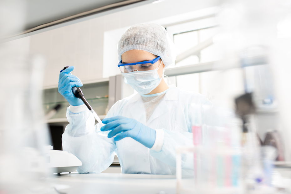 A laboratory technician wearing a white lab coat, gloves, a face mask, and protective goggles is using a pipette to transfer liquid in a lab setting. The background shows lab equipment and glassware.