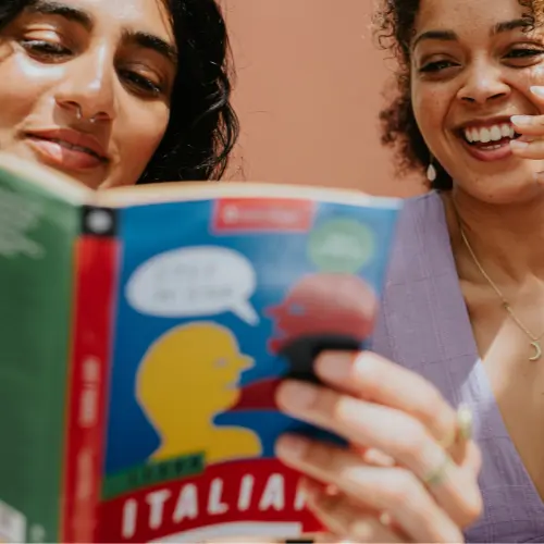Zwei Frauen sitzen zusammen und schauen freundlich auf ein Buch, das sie gemeinsam lesen. Das Buchcover zeigt Silhouetten von zwei Gesichtern in Gesprächen. Eine der Frauen lächelt, während die andere konzentriert liest. Sie genießen offensichtlich den Moment des gemeinsamen Lesens.