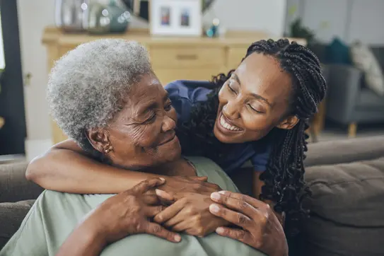 A young woman smiles while embracing an elderly woman in a cozy living room setting. They share a joyful moment, highlighting a strong bond between them. The background features soft furnishings and decor, creating a warm atmosphere.