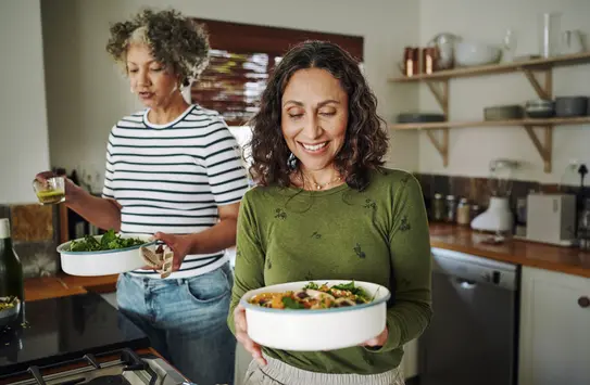 Two women are in a kitchen. One woman, with curly hair, holds a bowl of salad and smiles, while the other, wearing a striped shirt, pours a drink from a glass. The kitchen has modern appliances and a warm, inviting atmosphere.
