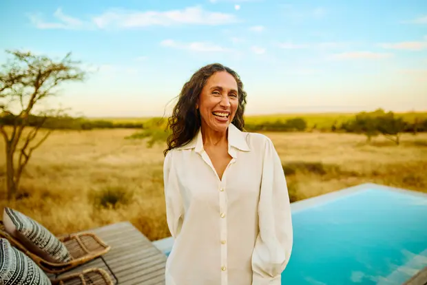 A smiling woman stands outdoors near a pool, surrounded by a grassy landscape and trees under a blue sky. She wears a light-colored, loose-fitting shirt, radiating warmth and joy.