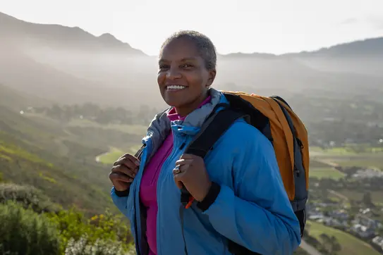 A smiling woman stands outdoors against a scenic backdrop of mountains and valleys. She wears a blue jacket and has a backpack, suggesting she is on a hike or outdoor adventure. The sun casts a warm glow, enhancing the natural beauty of the setting.
