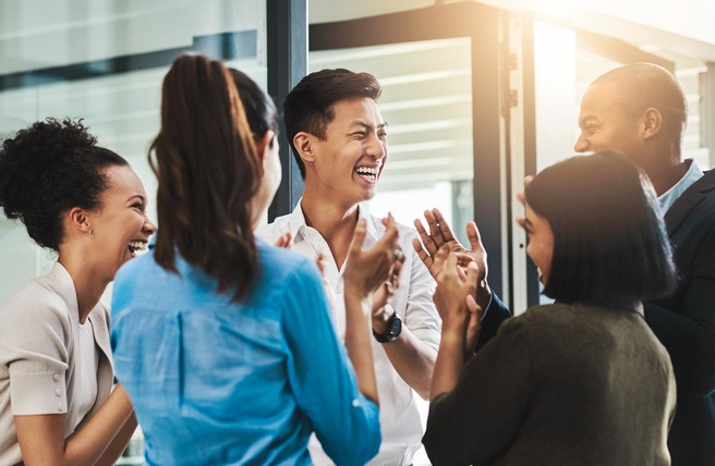 A group of five people, consisting of two men and three women, are laughing and engaging joyfully in a bright, modern office environment. They appear to be celebrating or sharing a lighthearted moment together.