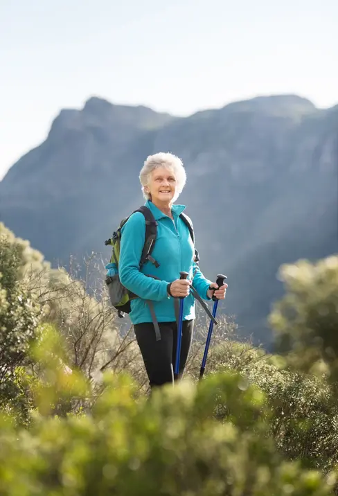 An older woman with short gray hair stands outdoors, smiling while holding walking sticks. She wears a blue jacket and a backpack, with a scenic mountain landscape in the background.