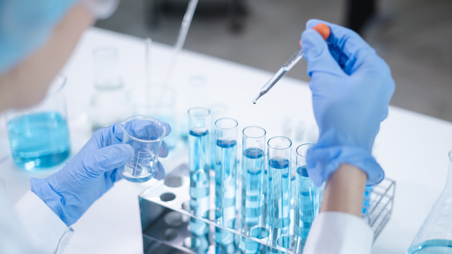 A laboratory setting with a scientist wearing gloves and a lab coat. The scientist is holding a small beaker in one hand and using a dropper to add liquid to a row of test tubes filled with blue liquid.