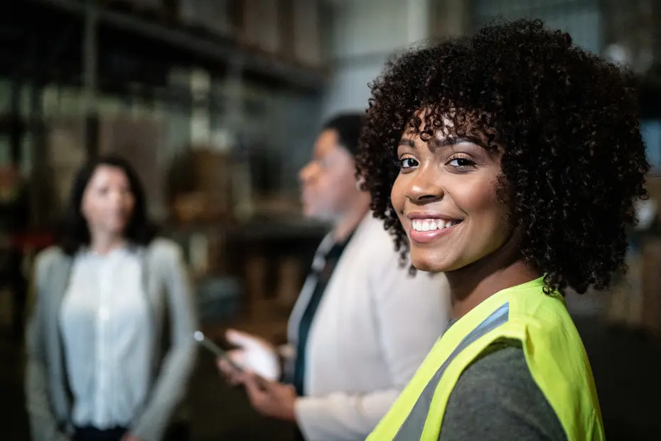 A smiling woman wearing a yellow safety vest stands in a warehouse, looking at the camera. Two other women are blurred in the background, engaged in conversation. The setting appears to be a work environment, with shelves and boxes visible.