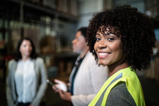 A smiling woman wearing a yellow safety vest stands in a warehouse, looking at the camera. Two other women are blurred in the background, engaged in conversation. The setting appears to be a work environment, with shelves and boxes visible.