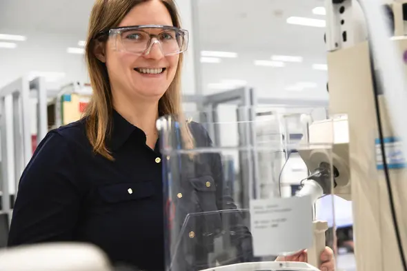 A smiling woman wearing safety glasses stands beside a laboratory machine. She is dressed in a black shirt and appears engaged in a scientific task. The background features laboratory equipment and a bright, spacious environment.