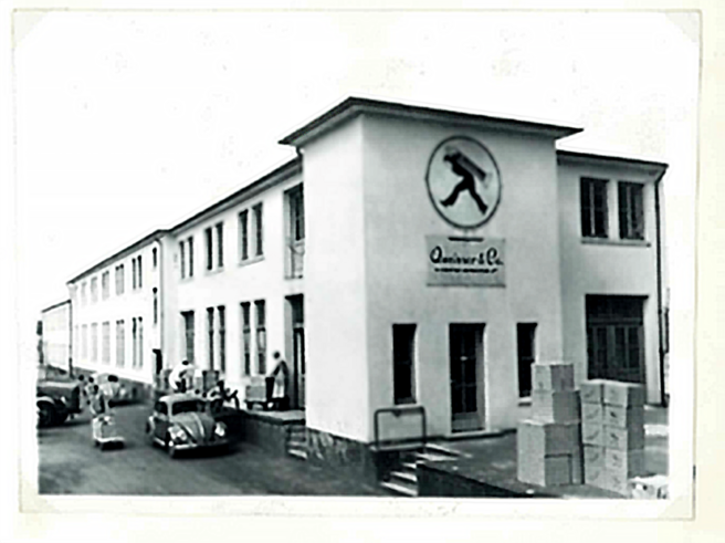 A black and white photograph of a large building with a logo featuring a person walking on its facade. There are several people gathered at the entrance, and various boxes are stacked nearby. Classic cars are parked in front of the building.