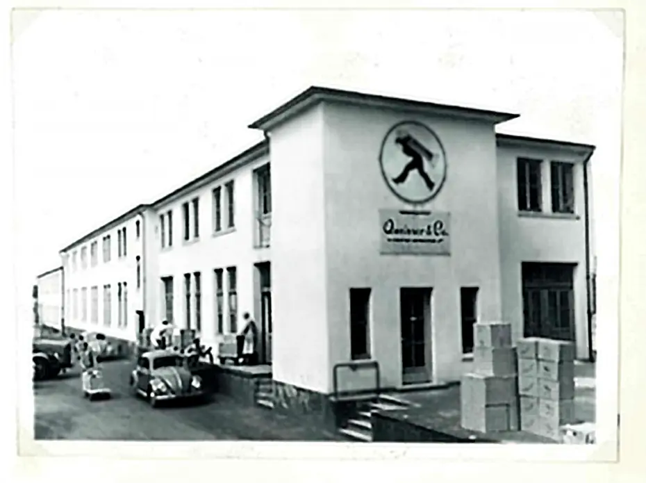 A black and white photograph of a large building with a logo featuring a person walking on its facade. There are several people gathered at the entrance, and various boxes are stacked nearby. Classic cars are parked in front of the building.