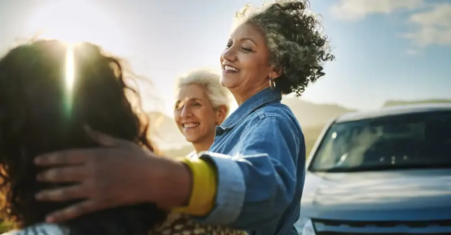 Three women share a joyful moment outdoors. One woman embraces another, while a third woman smiles nearby. The setting appears sunny, with a car parked in the background. The scene conveys warmth and connection among friends.