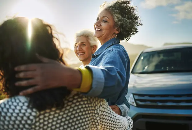 Three women share a joyful moment outdoors. One woman embraces another, while a third woman smiles nearby. The setting appears sunny, with a car parked in the background. The scene conveys warmth and connection among friends.