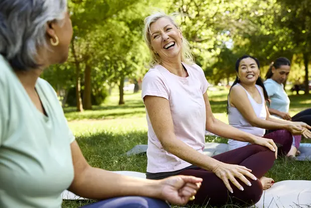 Four women sit on the grass in a park, engaging in conversation and laughing. They appear relaxed, wearing casual athletic clothing. The background features lush green trees and a sunny environment, creating a cheerful and inviting atmosphere for outdoor activities.