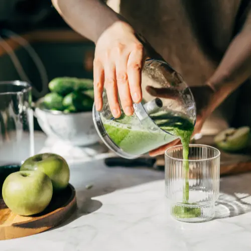 Eine Hand gießt einen grünen Smoothie aus einem Mixer in ein Glas. Auf dem Tisch liegen grüne Äpfel und Gurken, während Sonnenlicht auf die Szene scheint.