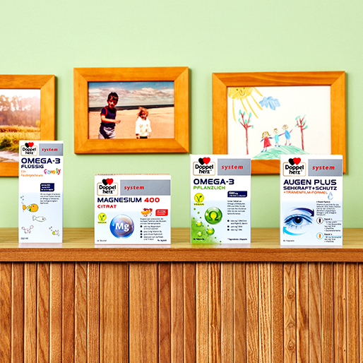 A wooden shelf displays various health supplement boxes, including "Omega-3 Flüssig," "Magnesium 400 Citrate," and "Augen Plus." Behind the shelf, a green wall features framed family photos and children's drawings.