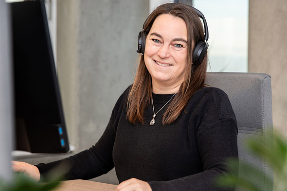A woman with long brown hair wearing headphones smiles while sitting at a desk with a computer monitor. She is dressed in a black sweater and appears friendly and engaged. Green plants are partially visible in the foreground.