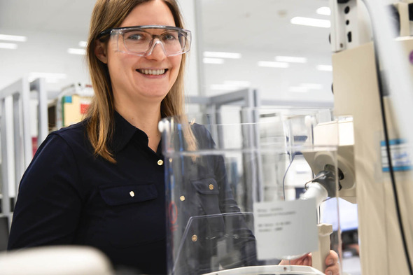 A smiling woman wearing safety glasses stands beside a laboratory machine. She is dressed in a black shirt and appears engaged in a scientific task. The background features laboratory equipment and a bright, spacious environment.