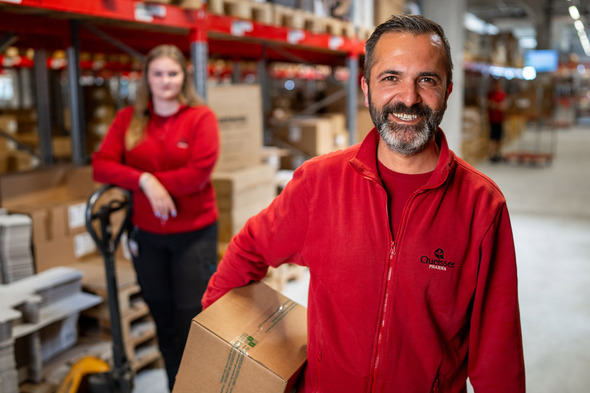 A smiling man in a red jacket holds a box in a warehouse setting, with shelves stocked behind him. A woman in a similar red jacket stands in the background, leaning against a stack of boxes, with a pallet jack nearby. The scene conveys a productive work environment.