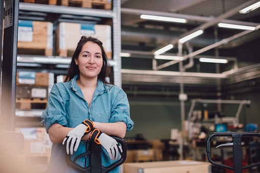 A smiling woman in a blue shirt and gloves stands next to a hand pallet truck in a warehouse. The background shows shelves with boxes and various equipment, indicating a busy work environment.